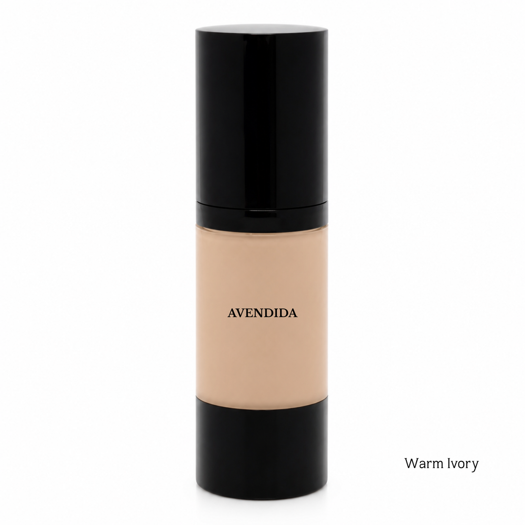 A bottle of AVENDIDA liquid foundation in the shade Warm Ivory, featuring a glossy black cap and base with a beige-toned center showing the liquid makeup. The brand name 'AVENDIDA' is printed in black on the beige section, and 'Warm Ivory' is written in small text in the bottom right corner against a solid white background.