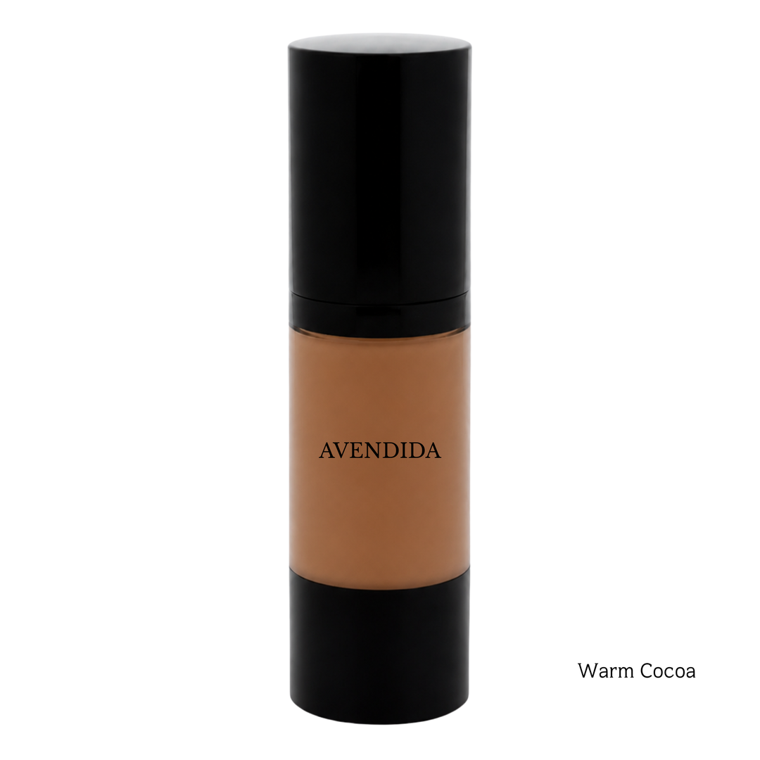 A bottle of AVENDIDA liquid foundation in the shade Warm Cocoa, featuring a warm brown liquid visible through a clear cylindrical bottle with a glossy black cap and base, set against a plain white background.
