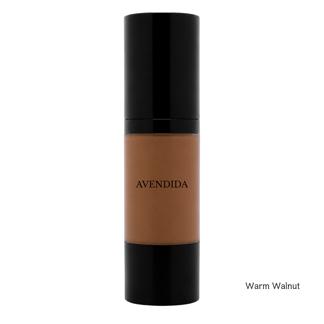 A bottle of AVENDIDA liquid foundation in the shade Warm Walnut, featuring a warm brown liquid visible through a clear cylindrical bottle with a glossy black cap and base, set against a plain white background.