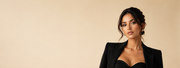 A woman with dark hair styled in an updo, wearing a black blazer over a black sweetheart neckline top, standing against a solid beige background with copy space on the left.