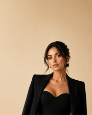A woman wearing a black structured blazer over a black sweetheart-neck top, photographed against a plain warm beige studio background.