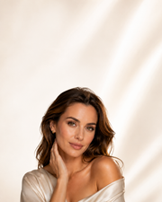 A woman with wavy brown hair wearing a champagne off-the-shoulder top, posing against a warm cream background with soft diagonal light shadows.